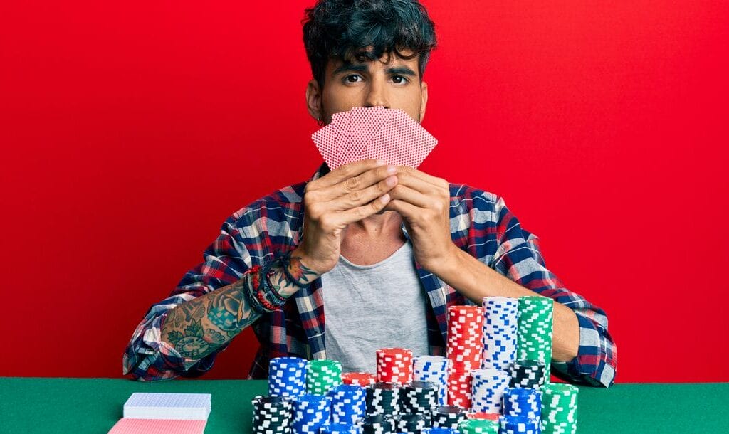 Young man playing poker and covering face with cards against a red background.