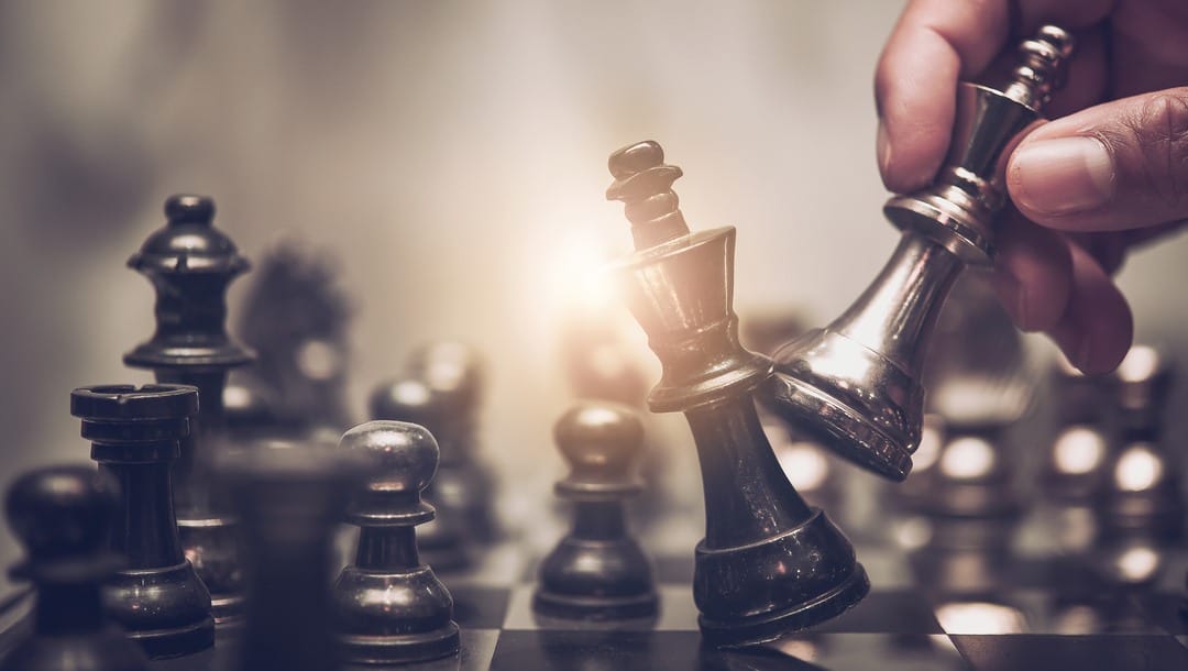 A hand holds a fallen king chess piece amid other chess pieces, representing defeat on the chessboard.
