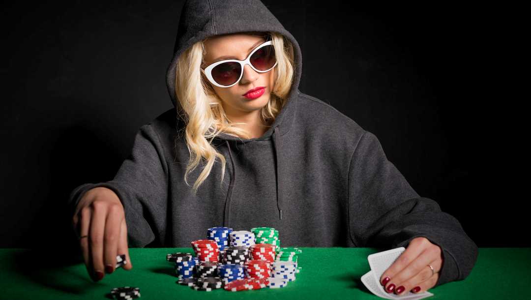 A woman wearing a hoodie and sunglasses checks her cards at a poker table.