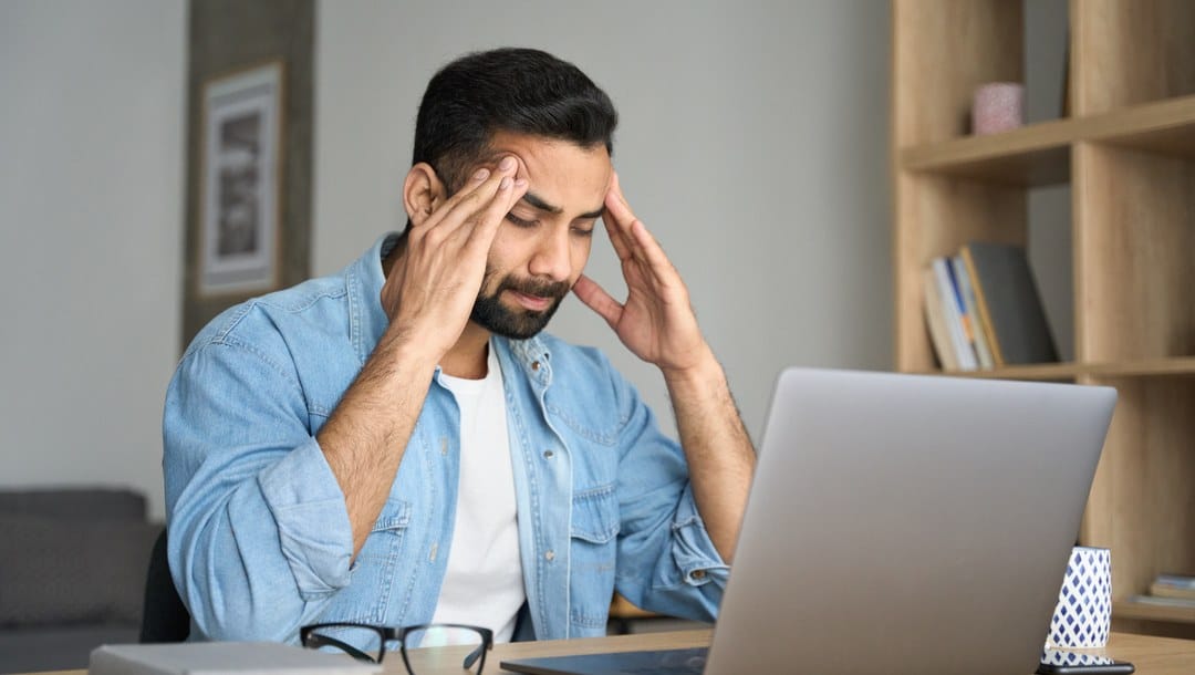 A stressed man sitting at his laptop rubs his temples.