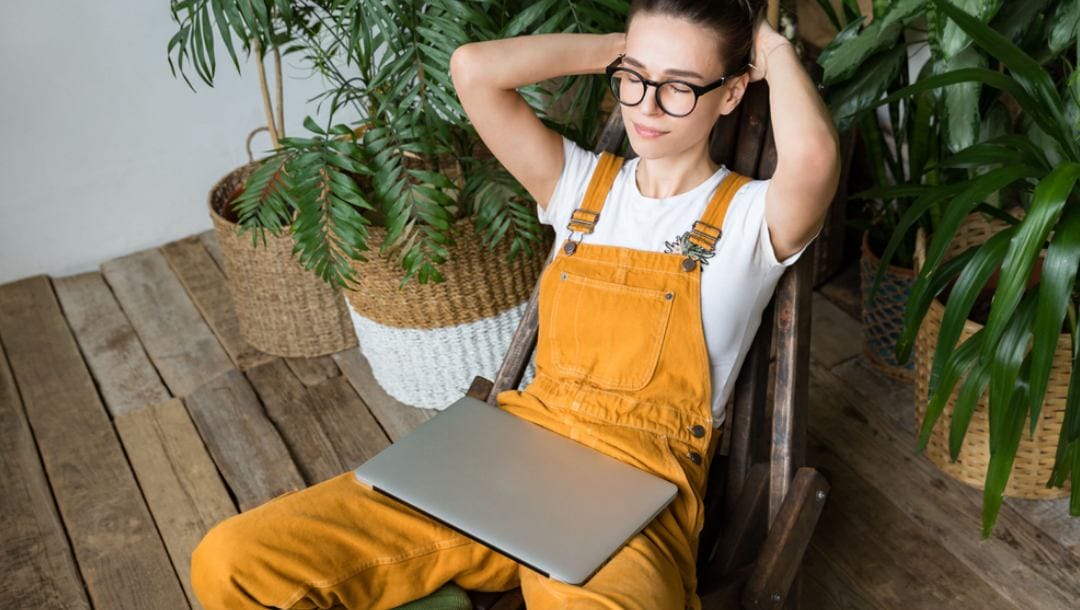 A woman in a chair leans back and closes her eyes with a closed laptop on her lap.