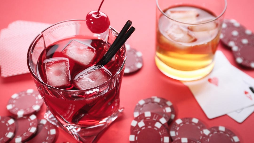A fruity pink cocktail and glass of whiskey surrounded by red casino chips and playing cards.