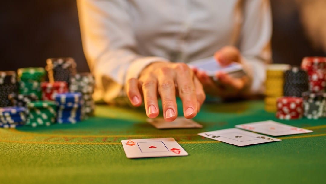 A poker game dealer holds one hand over a deck of cards while the other hand hovers above ace cards on a poker table.