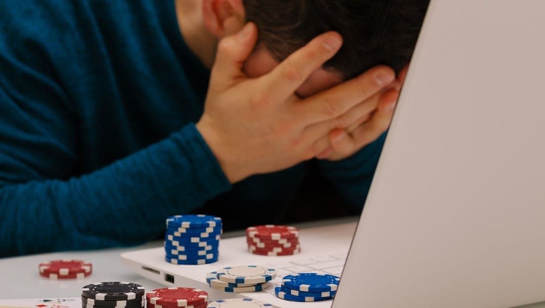 An online poker player holds his head in his hands while using his laptop. Poker chips sit next to and on his laptop.