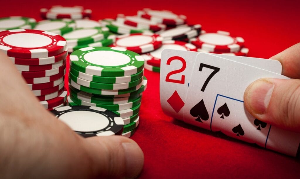A poker player shows a hand with 2 and 7 and holds some poker chips on a poker table.