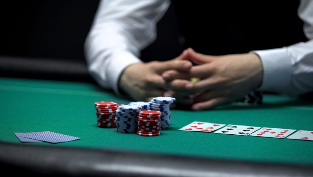 A person sitting with folded hands looking at cards and chips on a poker table.