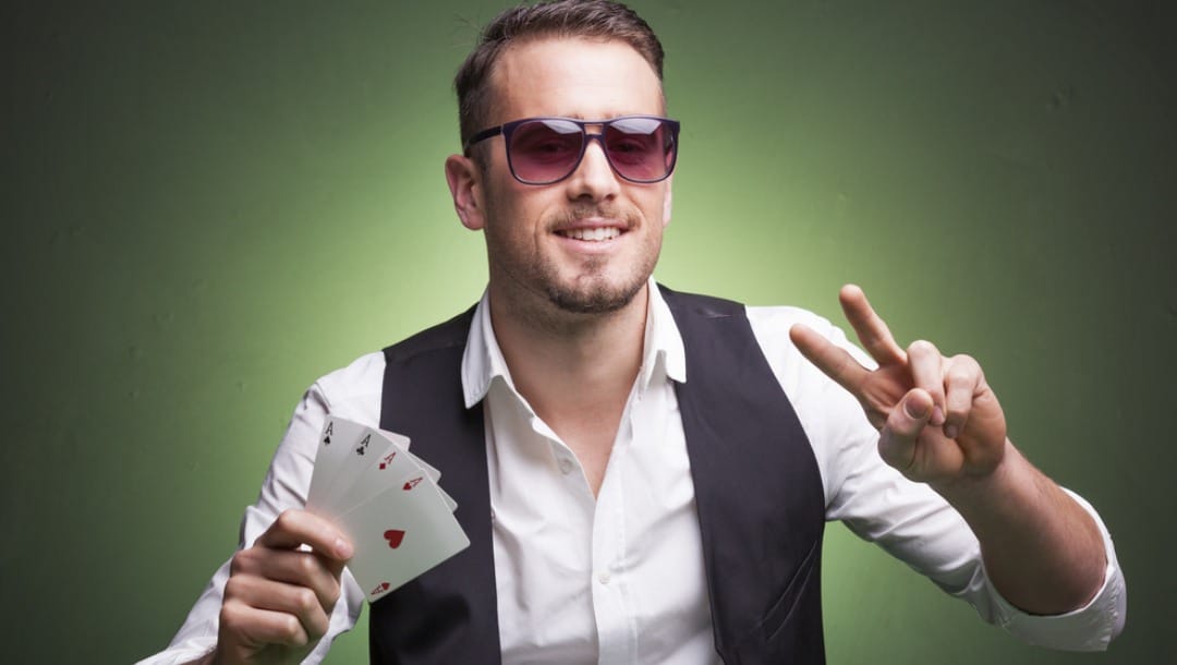 A poker player holds up a peace sign with his one hand and four aces in the other.