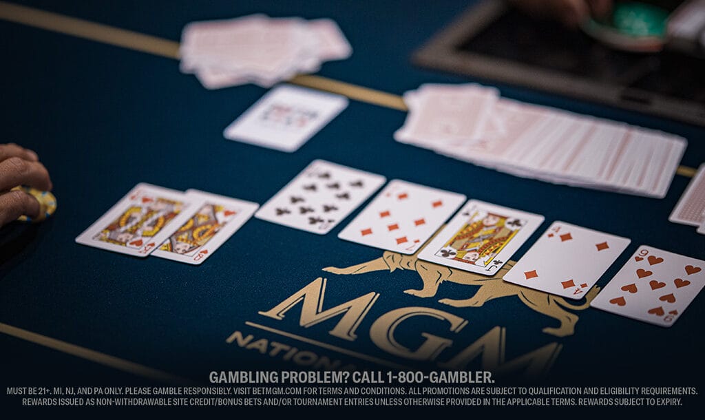 Poker game on a blue MGM table with playing cards laid out. Focus on a hand holding poker chips, conveying tension and strategy in gambling.
