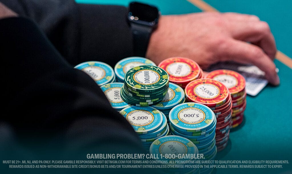 A close-up of poker chips stacked on a green table, with a person’s hand nearby. Chips in various colors, denominations visible, evoke a casino setting.