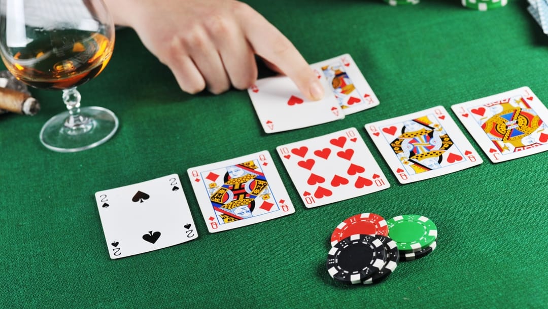 A gambling hand points to poker cards on a green felt table with poker chips and a glass of cognac.