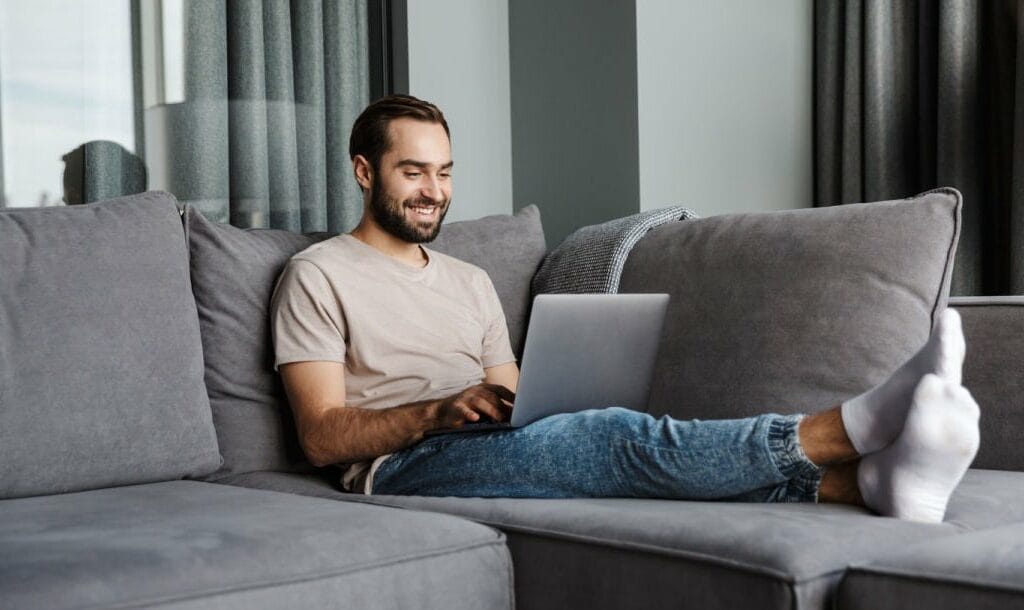 A man relaxing on a sofa looking at his laptop screen.