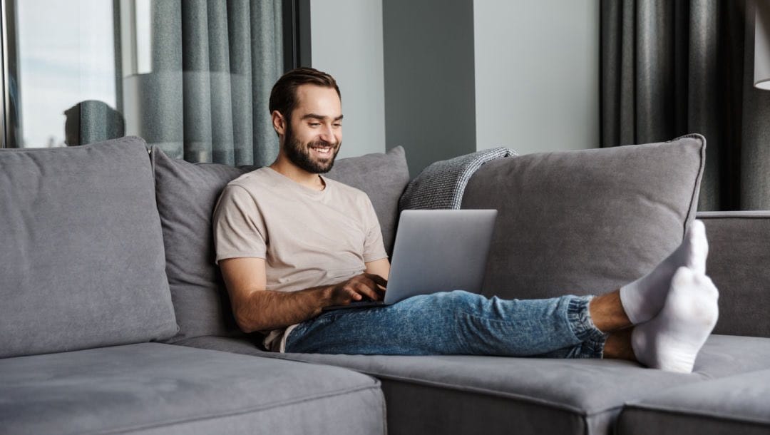 A man relaxing on a sofa looking at his laptop screen.