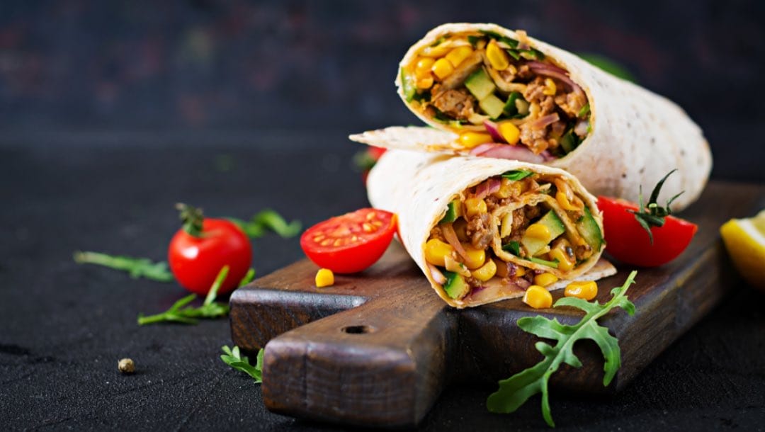 Two halves of a wrap filled with meat and vegetables on a wooden board with sliced cherry tomatoes and garnish around them.