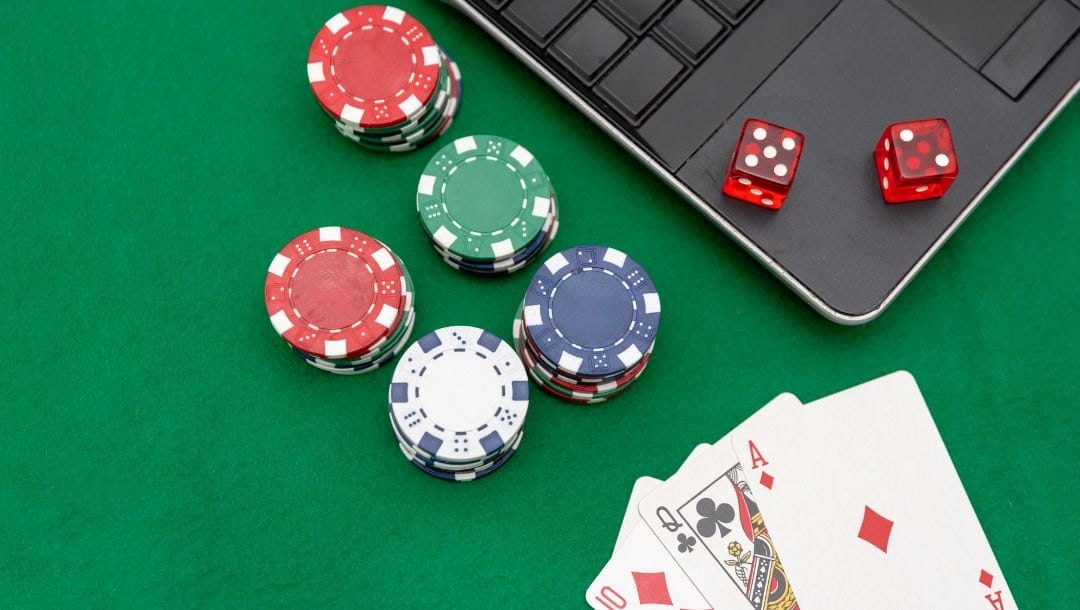 Stacks of poker chips, three playing cards, two red dice and a laptop arranged on a green poker table.