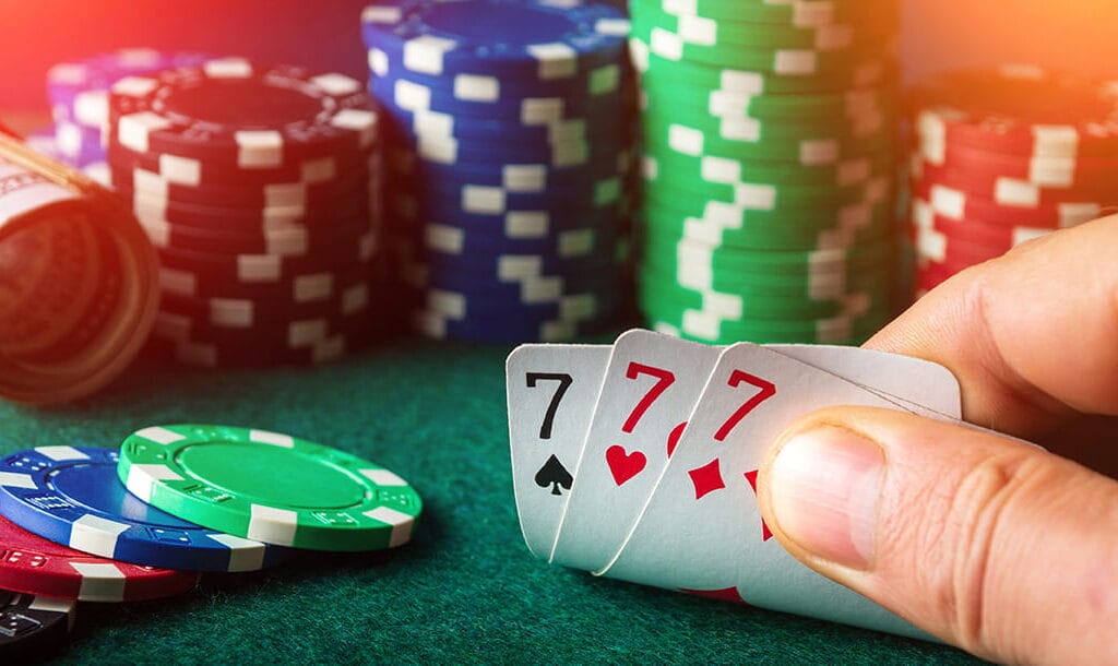 A person revealing their playing cards showing three 7’s, with stacks of poker chips and rolls of cash in the background.
