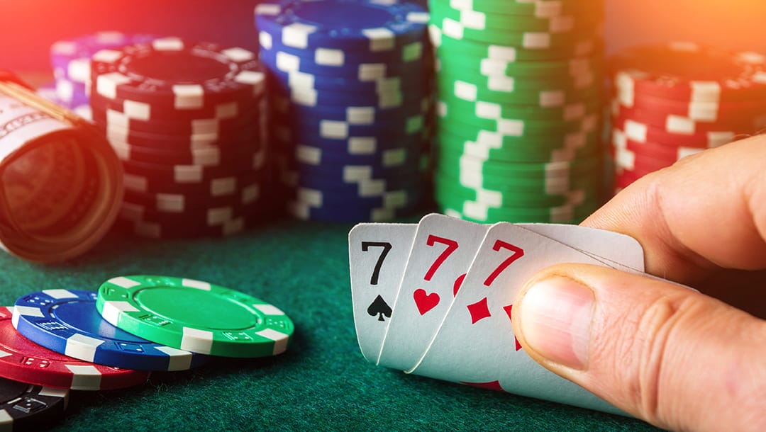 A person revealing their playing cards showing three 7’s, with stacks of poker chips and rolls of cash in the background.