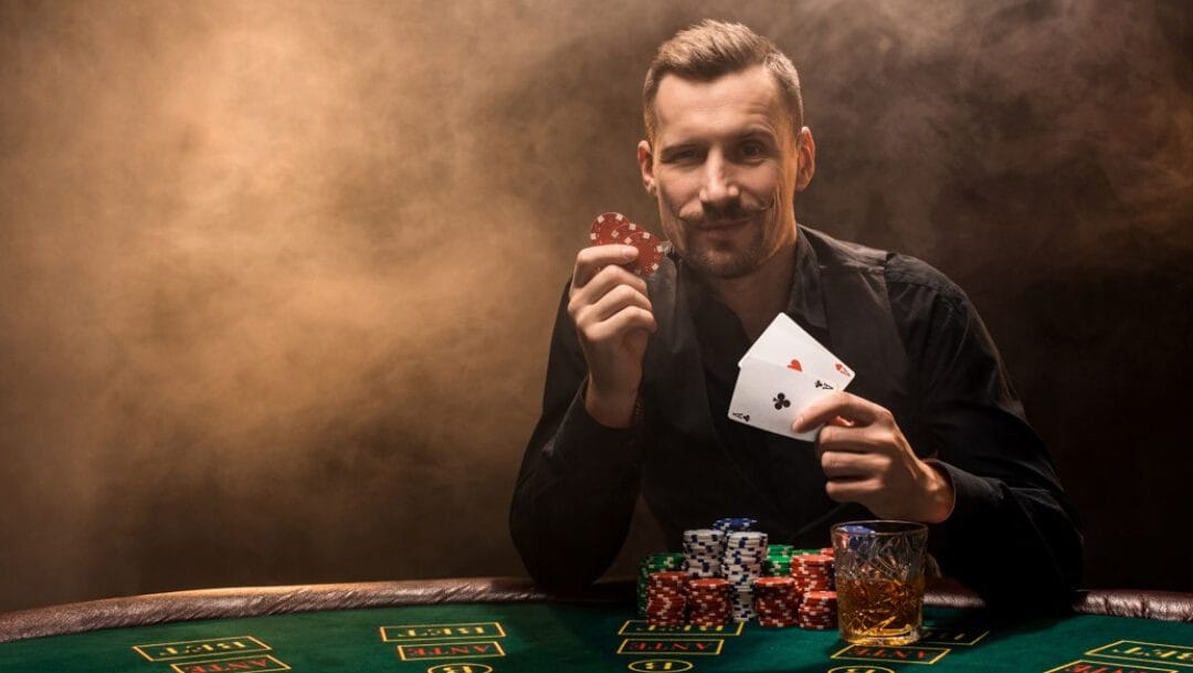 A man holds two aces and two poker chips, leaning on a poker table