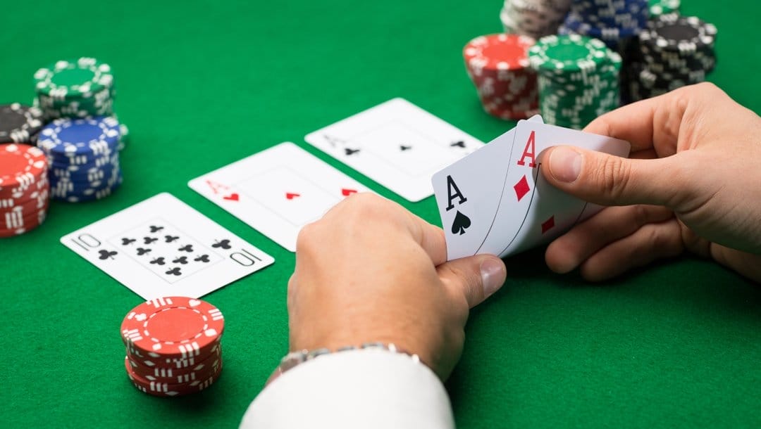 A poker player checks their hole cards in a game of Texas Hold’em. They see two aces. There are three community cards, including an additional two aces, as well as stacks of poker chips, on the table.