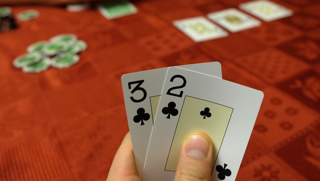 A poker player holds their hole cards, a two and a three of clubs.