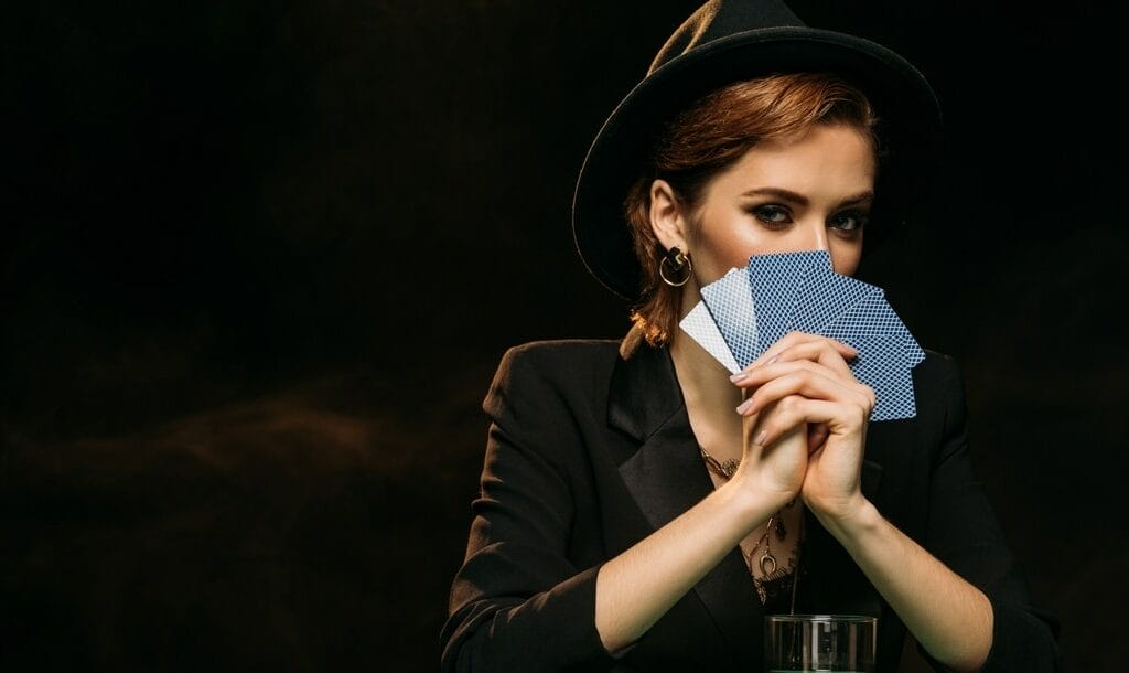 An elegantly dressed woman hides part of her face behind several playing cards.