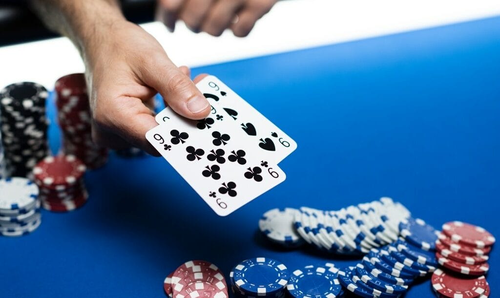 A poker player reveals two hole cards: a pair of nines. There are various stacks of chips in the background and the poker pot in the foreground.