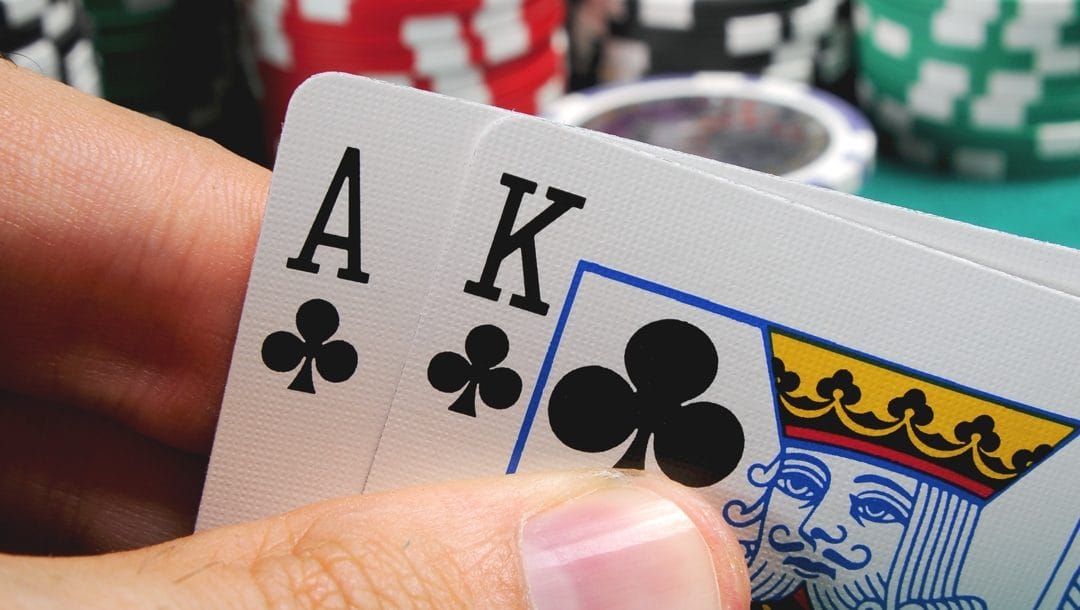A close-up of a poker player’s hand holding an ace and a king of clubs. There are stacks of poker chips in the background.