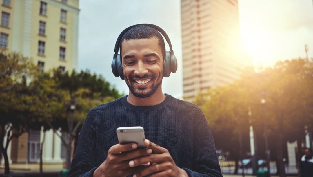 A man with headphones smiles as he looks at his smartphone.