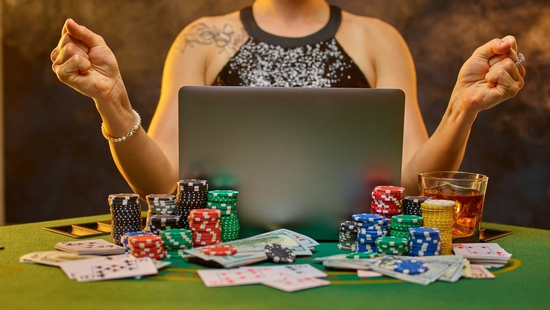 A woman clenches her fists in front of a laptop surrounded by poker chips, playing cards, a drink and cash.