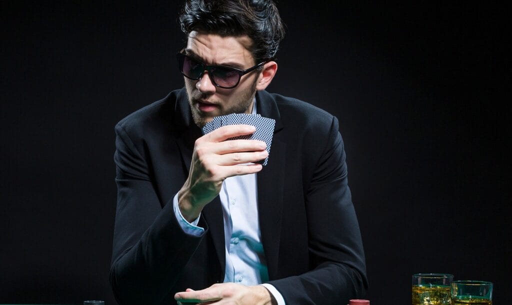 A person wearing a suit and sunglasses holds their poker hand up while looking to their left.