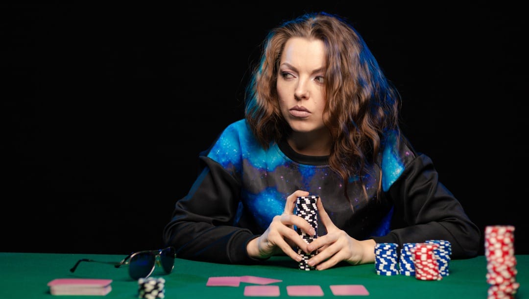 A person wearing a blue and black hoodie looks suspiciously to their left while holding a large stack of black-and-white poker chips.