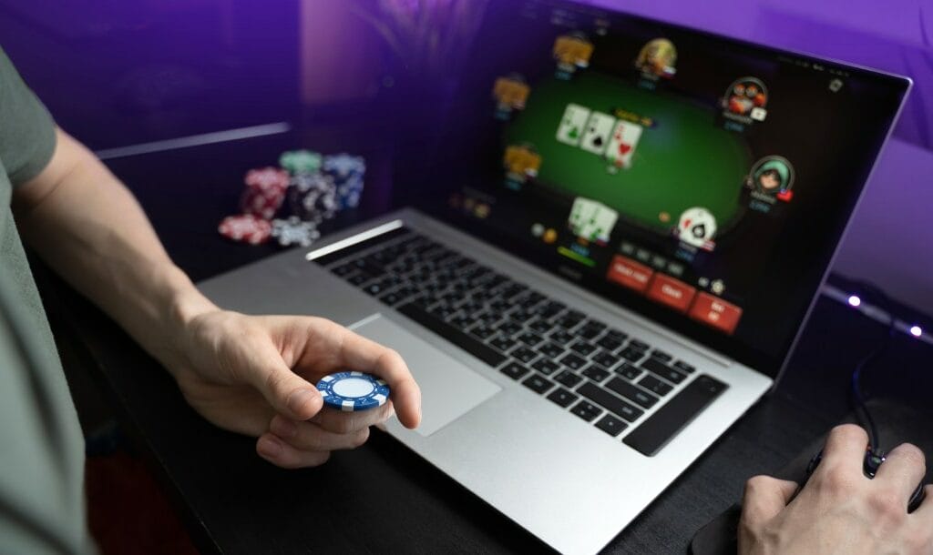 A man holding a casino chip while playing online poker on a laptop.