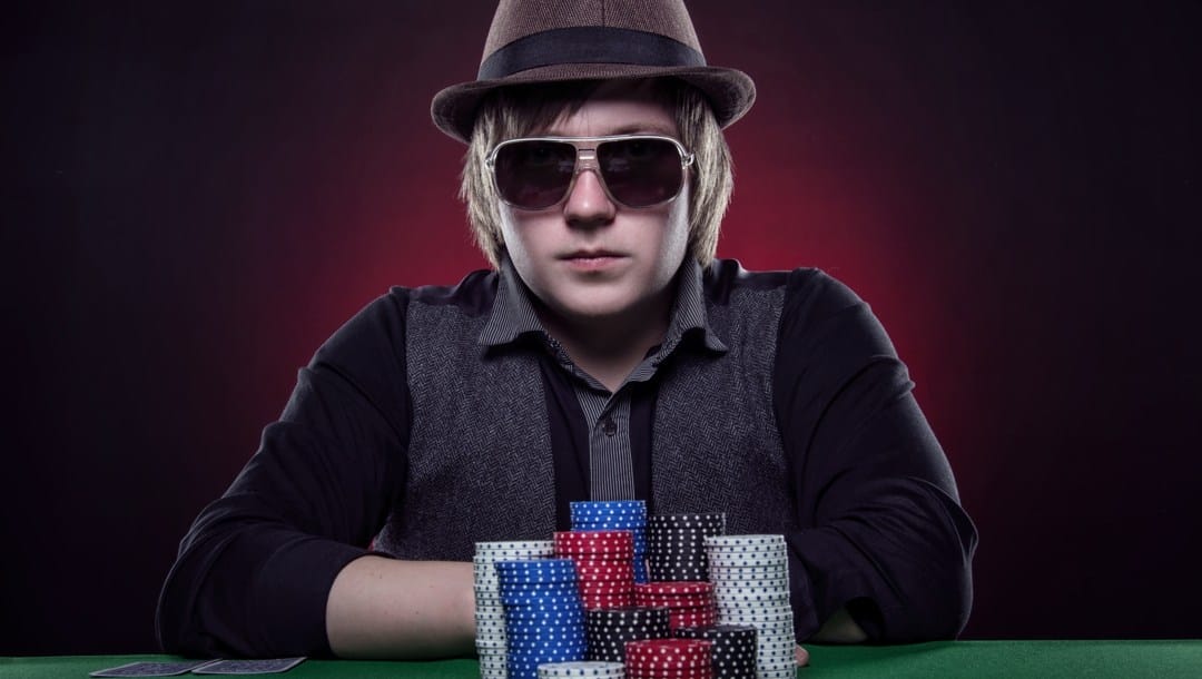 A man wearing sunglasses and a hat with an expressionless face sitting at a poker table. There are poker chips and two poker cards on the table.