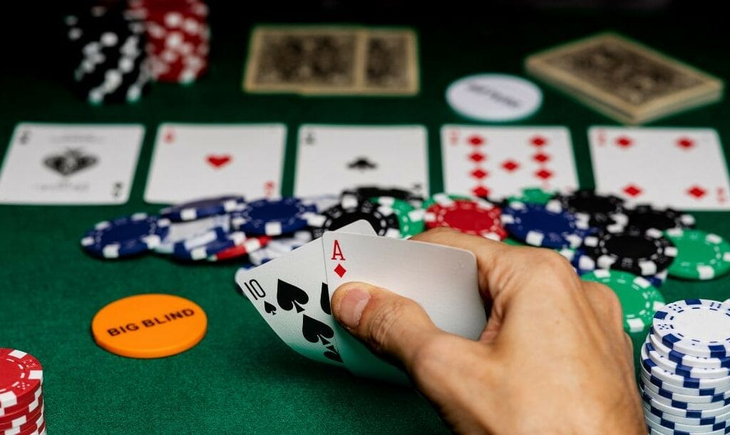 Player checking his hole cards of spade ten and diamond ace, in front of him on the poker table is a poker game comprising poker chips and playing cards post-flop