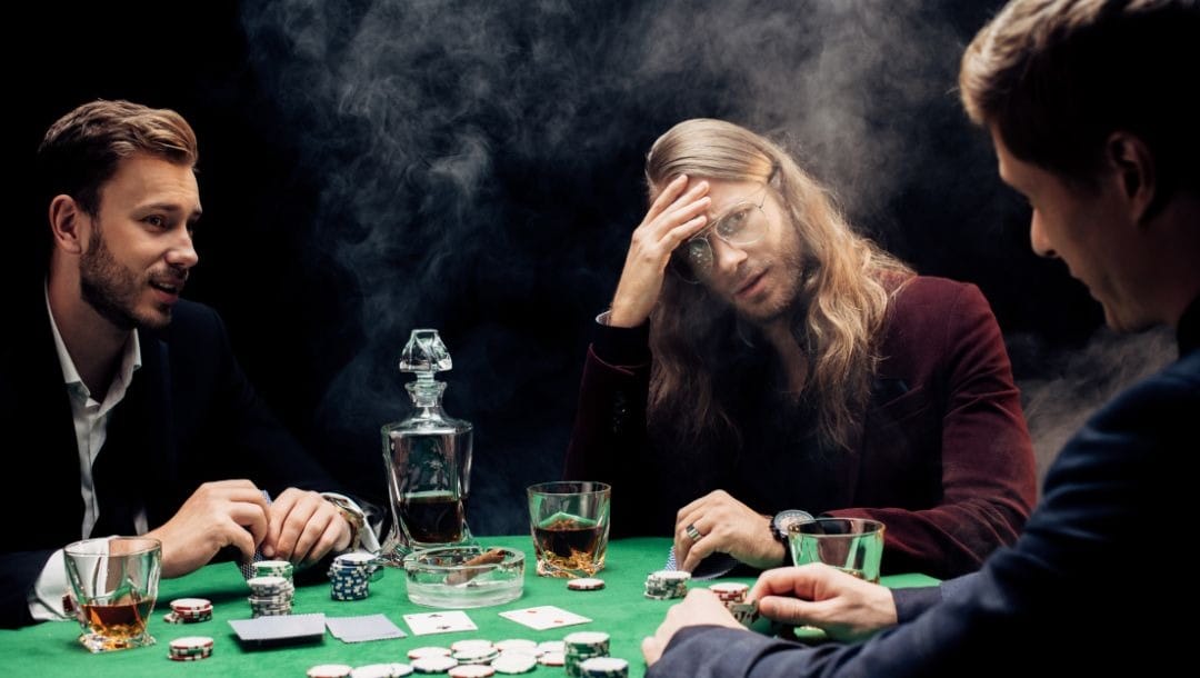 Three men sit around a table playing poker, one man checks his hole cards while the other two watch him, on the table are cards, poker chips and drinks