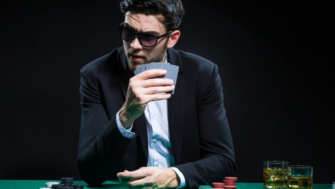 Man wearing sunglasses sitting at a poker table holding cards with poker chips in front of him and two drinks beside him on the table