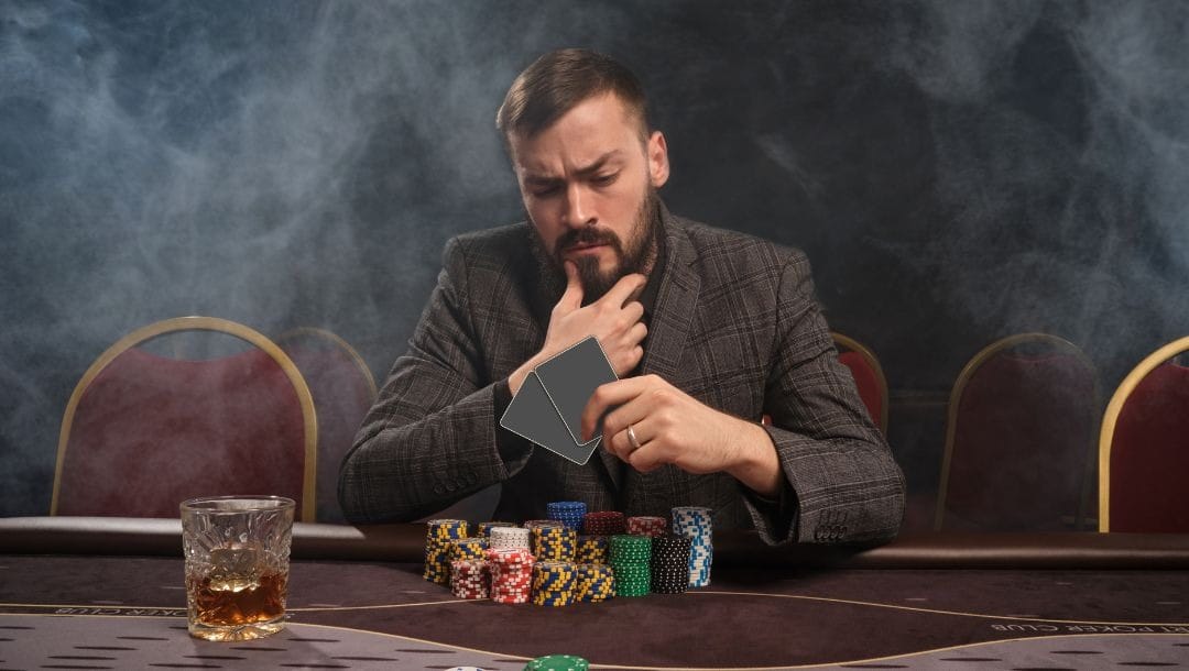 Man sitting at a poker table staring at his two cards in his hand with stacks of poker chips in front of him and a drink beside them, the air is smokey