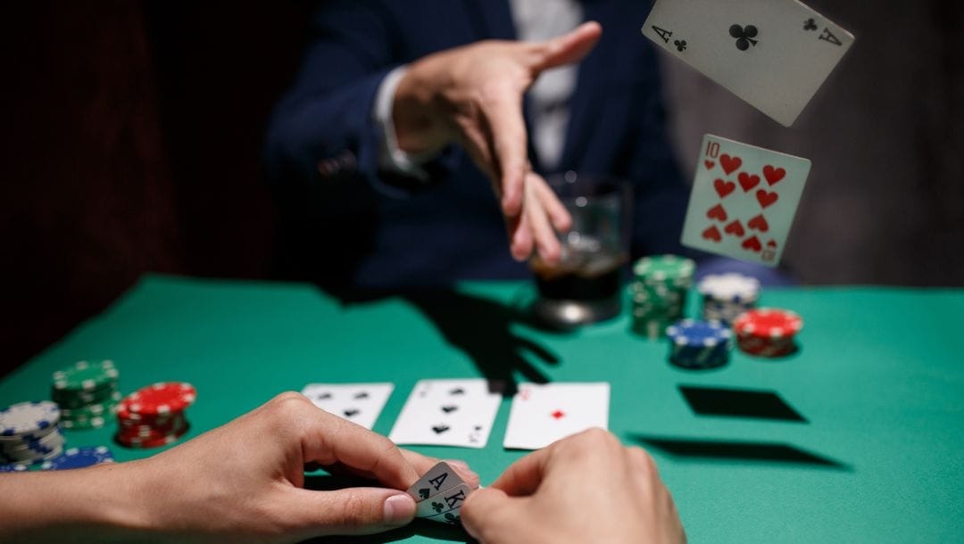 A pair of hands holds two cards in the foreground while a man folds in his poker hand in the background