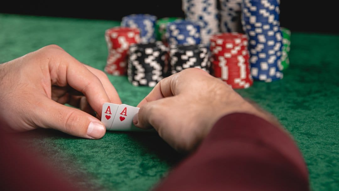 Player glimpsing at their cards on a green felt table, revealing two aces of hearts, with slightly blurred poker chip stacks in the background.