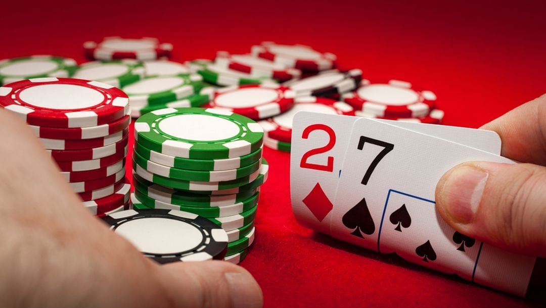 A poker player sitting at a casino table with poker chips glances at their two hole cards, revealing a 2 of diamonds and a 7 of spades.