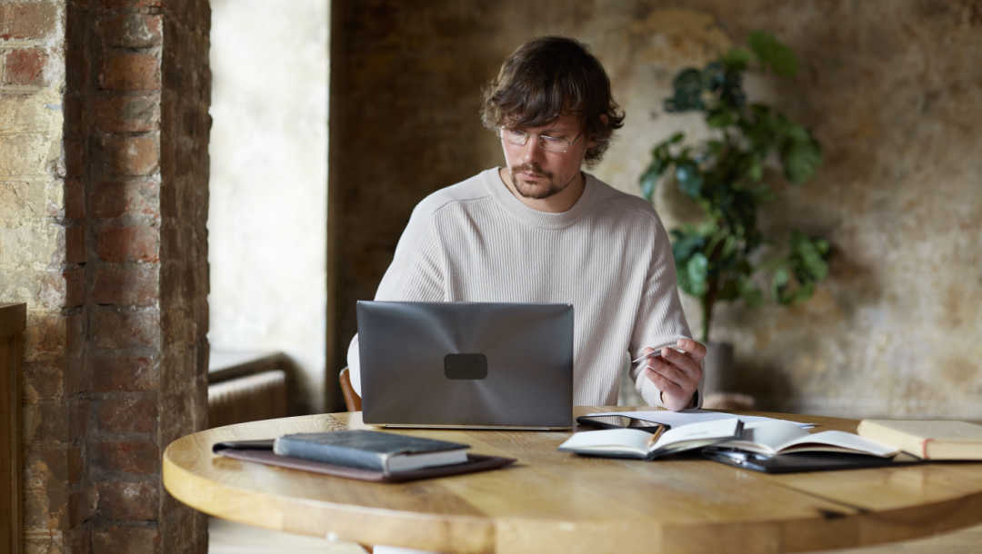 A man sitting at his laptop with books on the table next to him and a pen in his hand