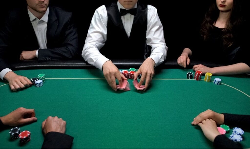 four poker players and a dealer, all in formal attire with poker chips stacked in front of them, sit around a green felt poker table as the dealer shuffles a deck of playing cards.