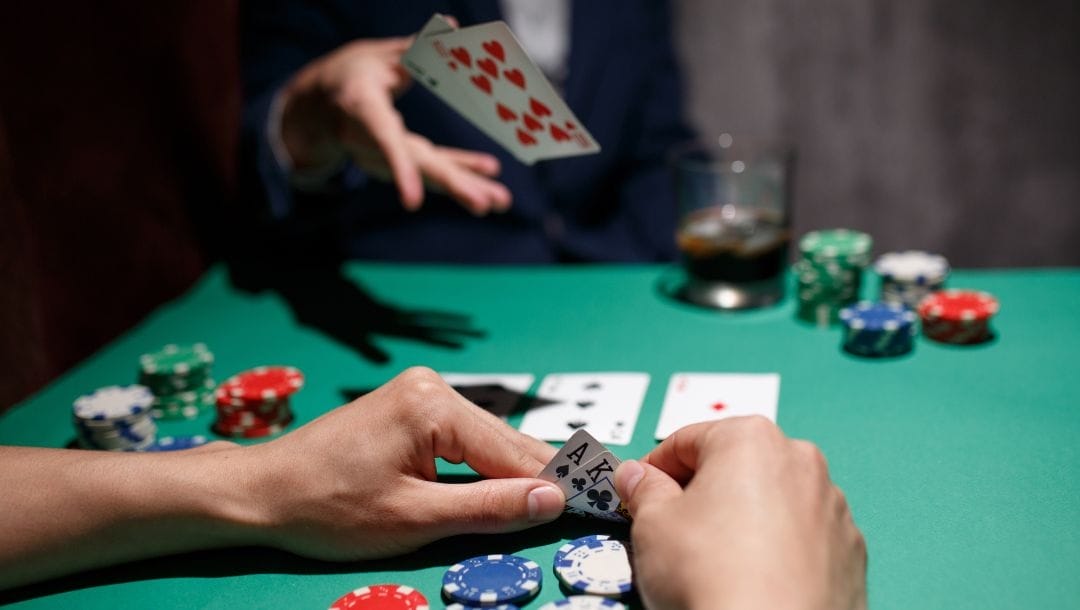 two players are sitting at a green felt poker table playing texas holdem, the player closest checks his hole cards of an ace and king while the player opposite him throws in his hole cards in defeat, on the table are poker chips, a drink and more playing cards
