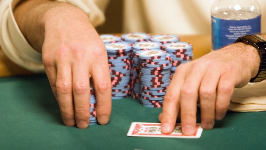 a close up of a man’s hands picking up a stack of poker chips with his right hand and touching his hole cards with his left hand that has a wrist watch on, in front of him are more stacks of poker chips and a bottle of water on a green felt poker table