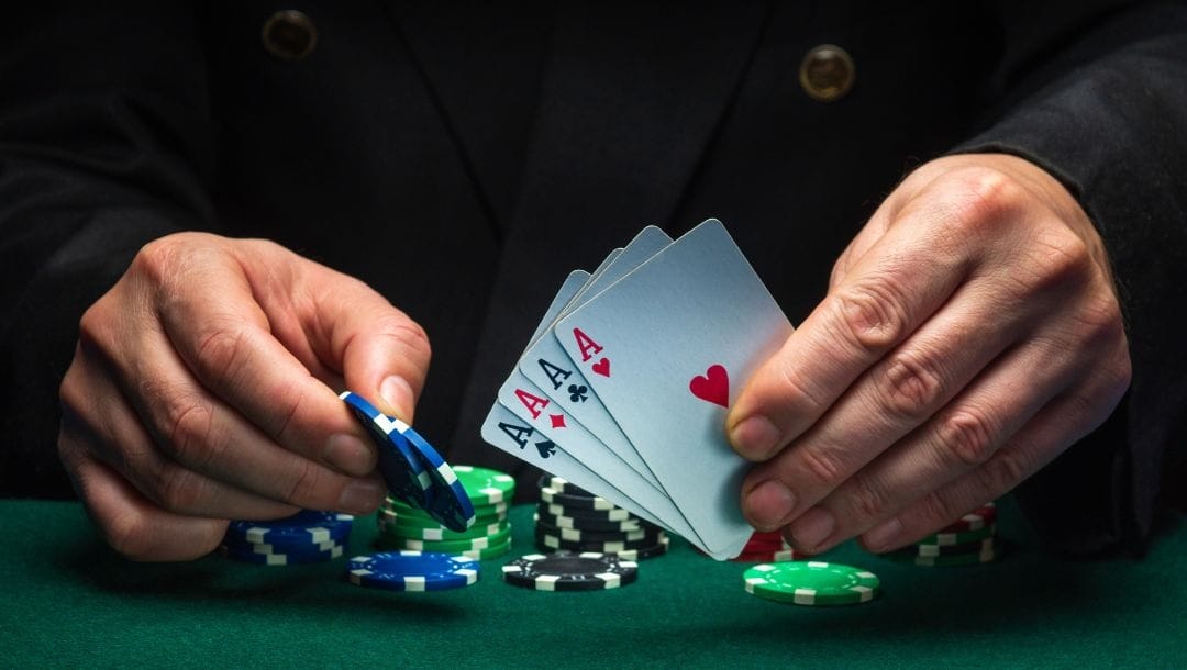 A man holding up four of a kind aces and two poker chips above a green felt poker table