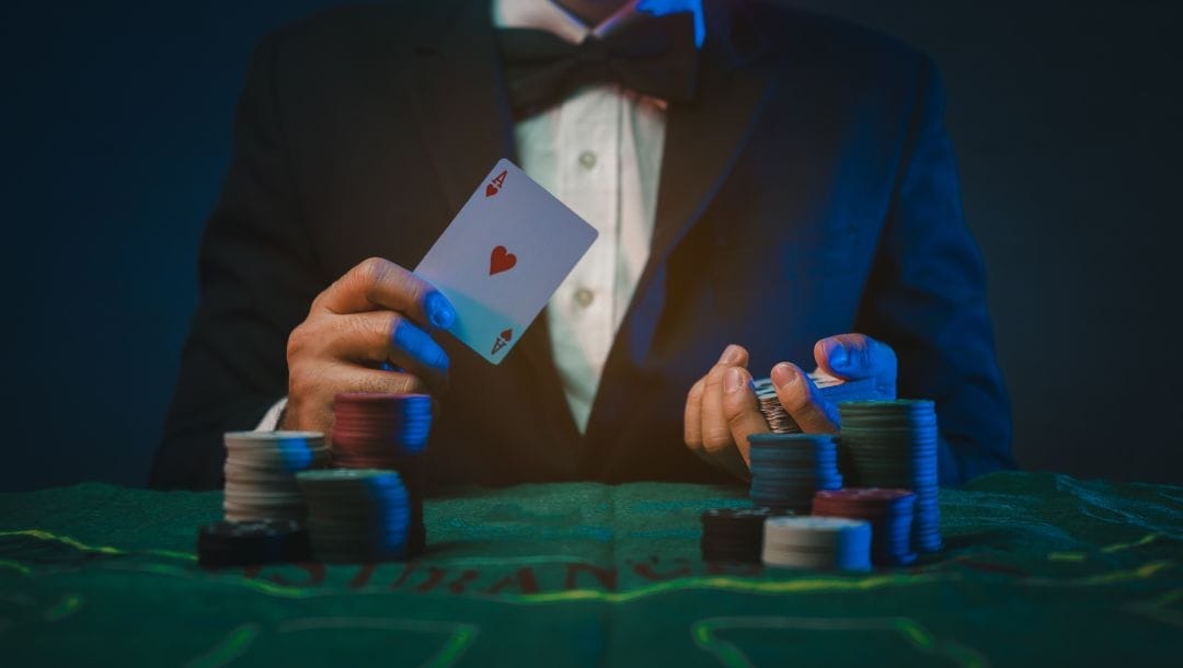 a man in a suit and bowtie sitting at a green felt poker table holding up an ace of hearts playing card with poker chips stacked in front of him