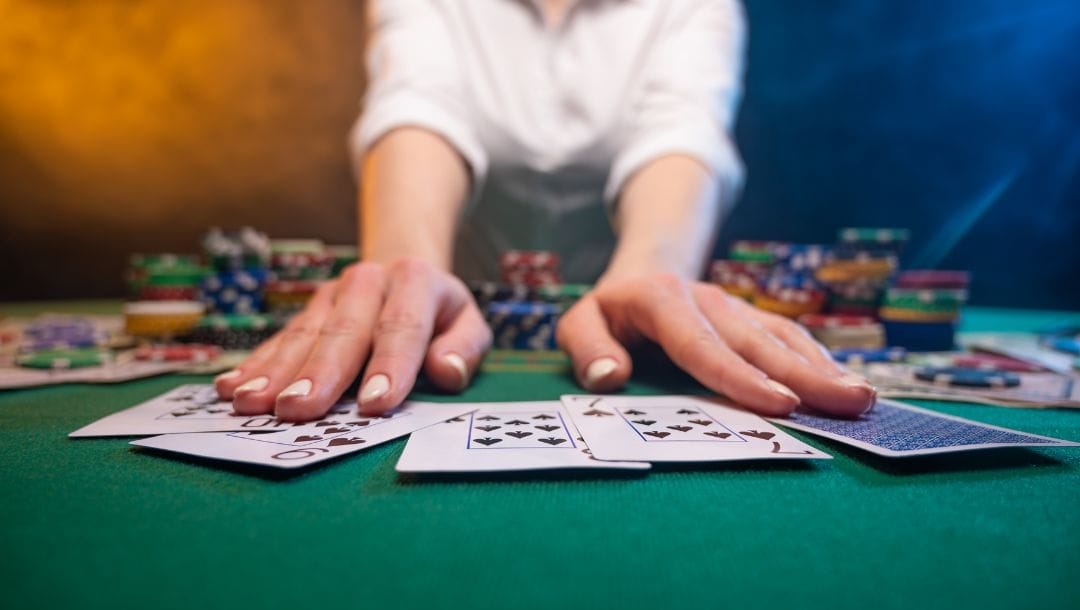 a woman with white painted nails pushes playing cards forward on a green poker surface with poker chips stacked in the background on the table