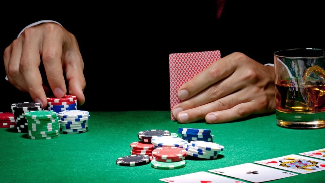 a man is picking up a stack of poker chips to place a bet during a poker match on a green felt poker surface, he holds his hole cards in his other hand beside his drink