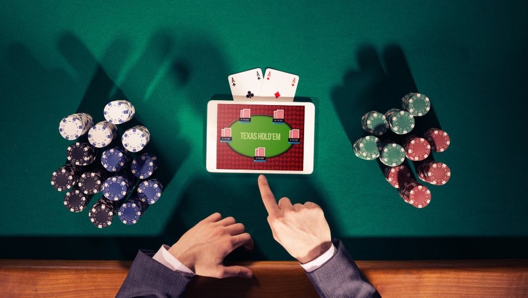 top view shot of a man sitting at a poker table with poker chips stacked on either side of him, in front of him is a digital tablet displaying an online Texas Hold’em poker game, a pair of ace playing cards stick out from underneath the top of the tablet
