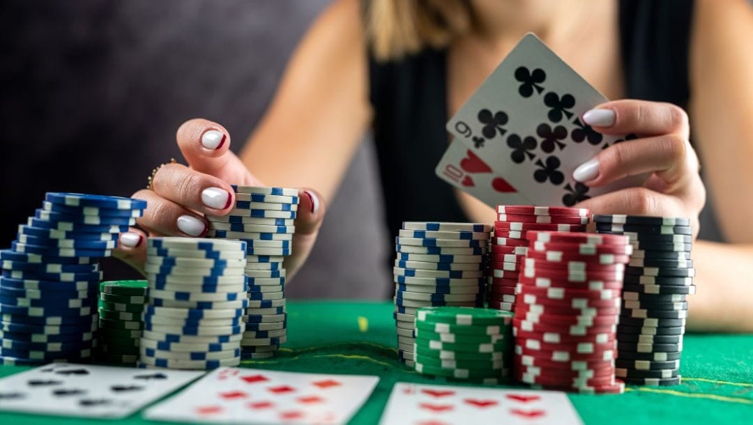 woman sitting at a green felt poker table holding two playing cards, nine clubs and ten hearts, in her left hand and touching one of several stacks of poker chips on the table