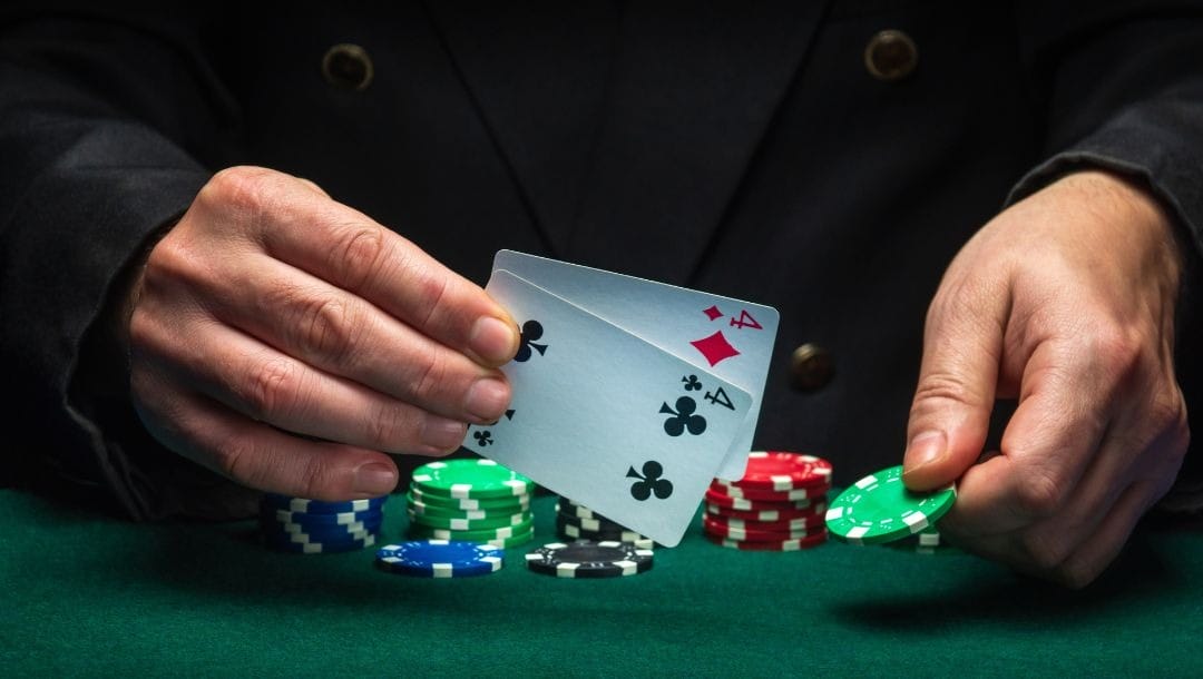 A man’s hands hold two playing cards in the right hand, a pocket pair of fours, and a green poker chip in the left hand above a green felt poker table with stacks of poker chips on it
